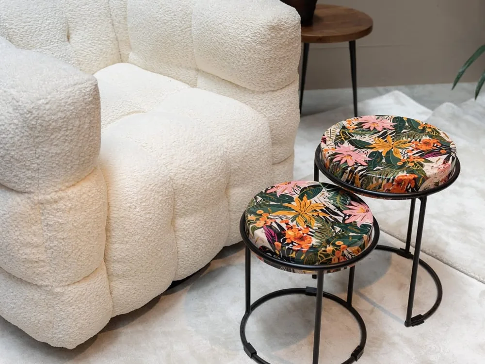 Deux tables d'appoint rondes à plateau imprimé de feuillages jaunes, verts et roses sur cercles en métal noir, devant un fauteuil en tissu bouclé écru